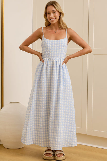 A woman modeling a light blue and white gingham maxi dress with spaghetti straps in a bright room with cream-colored wardrobes and a large mirror showing her reflection.