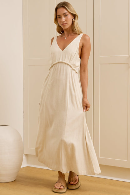A woman stands in a bright room wearing a sleeveless cream maxi dress with a V-neck and gathered waist. A large mirror on the left reflects her side profile, and two white ceramic vases sit on the floor against cream paneled walls.