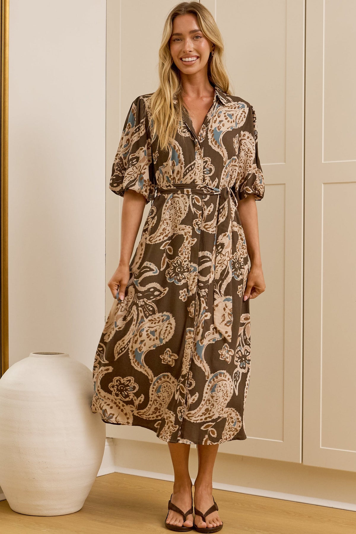 A woman stands in a bright room wearing a brown and beige paisley midi dress, with a large floor mirror on the left reflecting her side profile. The room has cream paneled walls and doors, light wood flooring, and a white ceramic vase on the floor.