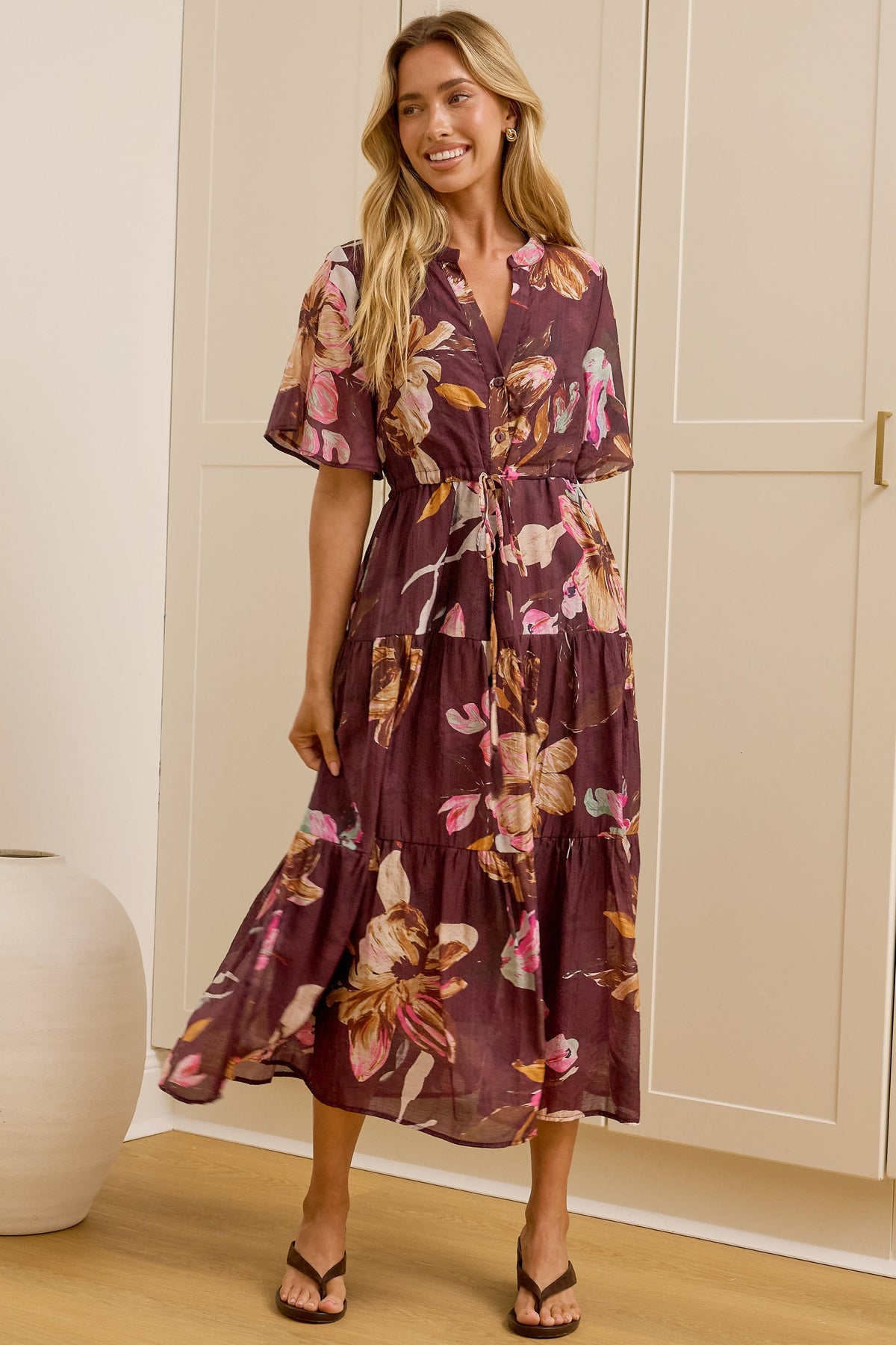 A woman models a purple floral midi wrap dress in a bright room with cream-colored wardrobes and a large mirror showing her profile. The dress features a purple base with pink, orange, and yellow floral patterns.