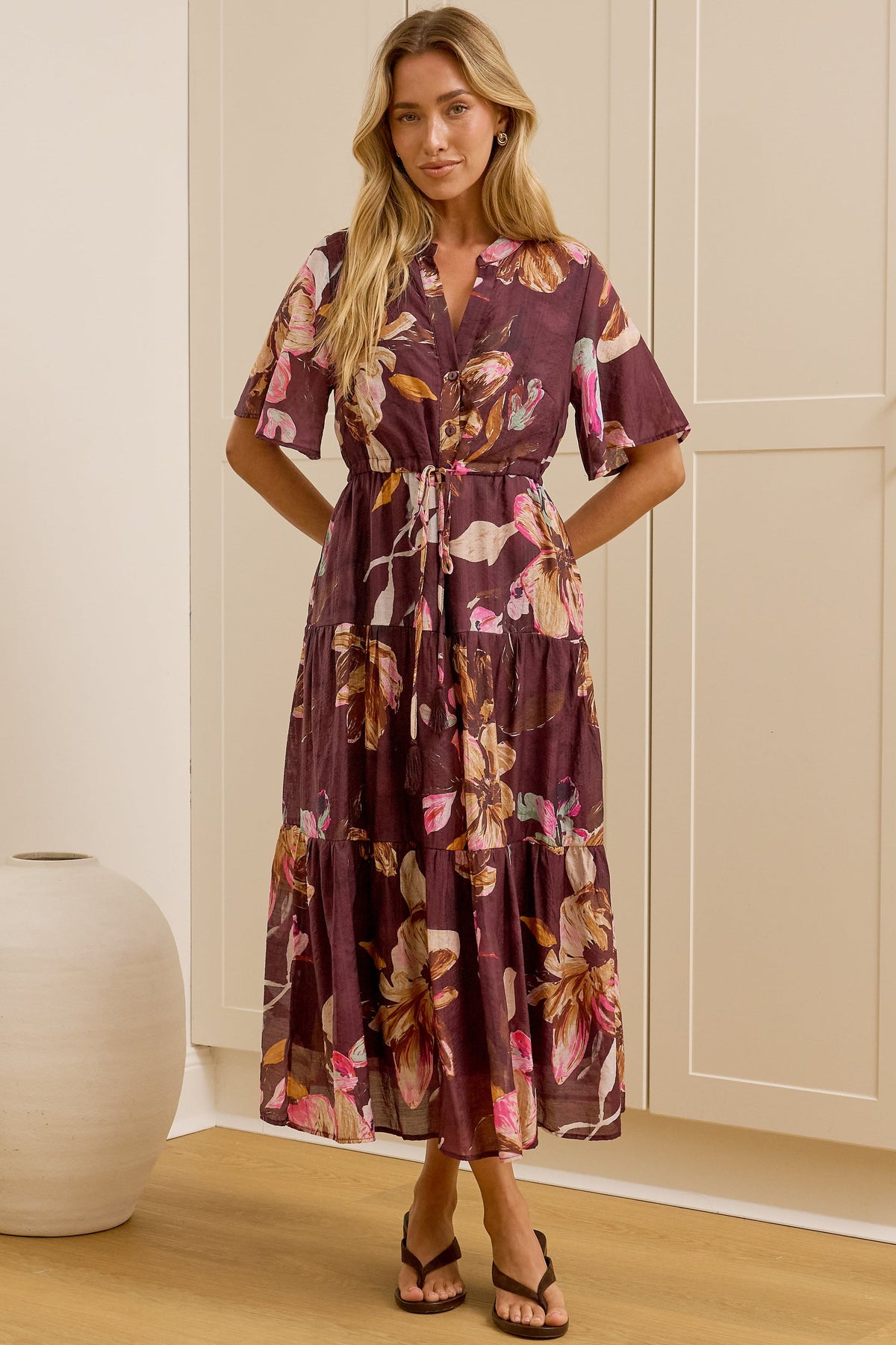 A woman stands in a bright room wearing a knee-length floral dress with short flutter sleeves and a tie waist. She is posed in front of a large mirror that reflects her side profile, with cream-colored cabinets and a light wood floor in the background.