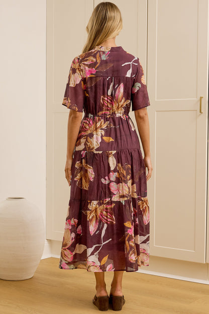 A woman stands with her back to the camera in a bright room, wearing a short-sleeved tiered maxi dress with a purple and pink floral print. She is positioned in front of white built-in wardrobes, with a large gold-framed mirror on the left reflecting her side profile.
