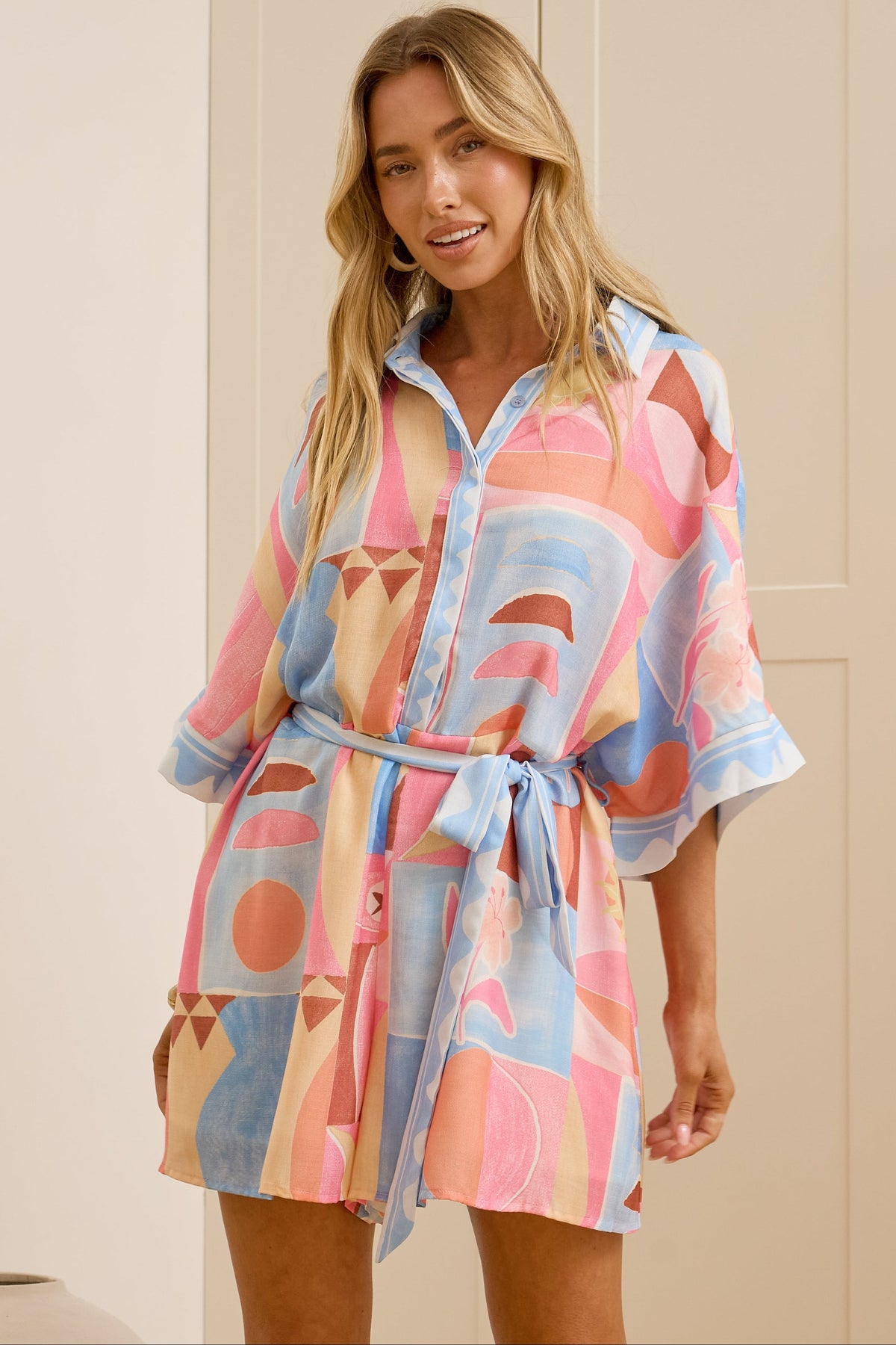 A woman modeling a short, button-front shirtdress with a tie waist, featuring a colorful abstract geometric print in pink, blue, and beige, set against a background of cream-colored paneled doors.
