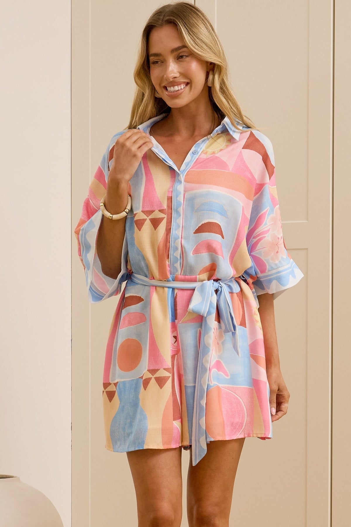 A woman stands in a bright room wearing a short, colorful abstract-patterned shirt dress with a tie waist. She is wearing beige slide sandals, and a large mirror on the left reflects her side profile.