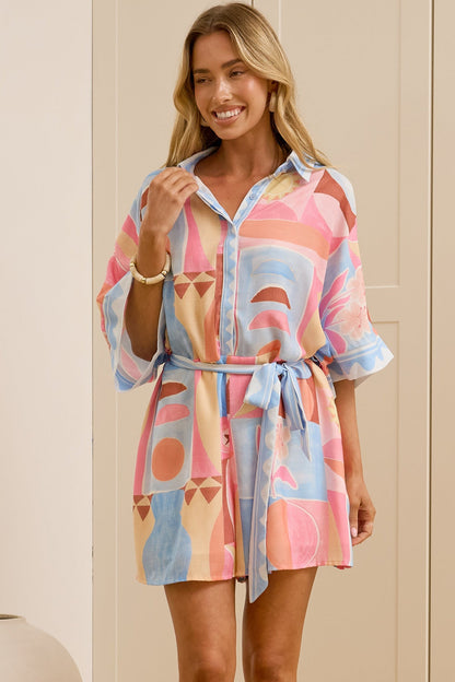 A woman stands in a bright room wearing a short, colorful abstract-patterned shirt dress with a tie waist. She is wearing beige slide sandals, and a large mirror on the left reflects her side profile.