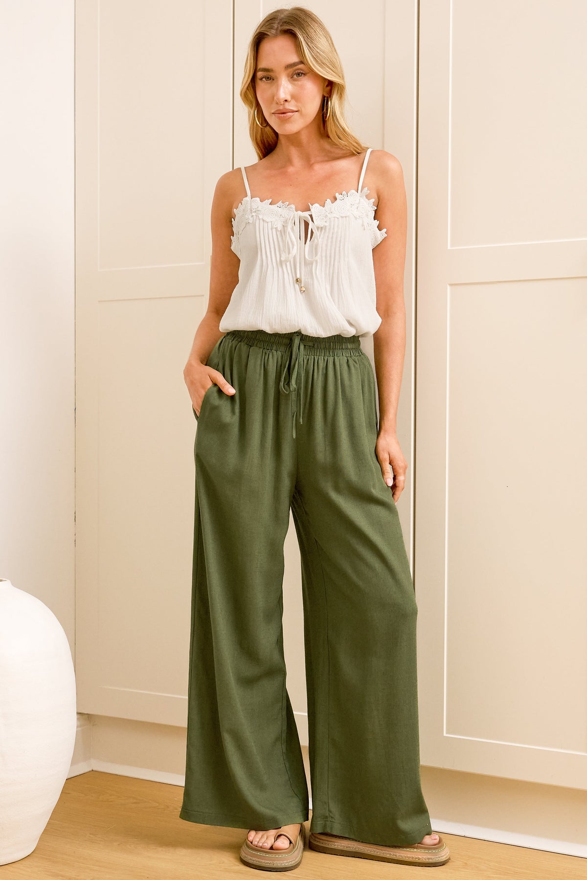 A woman wearing khaki linen pants with an elasticated waist, tie detail, and straight-leg fit, standing in front of a warm beige closet.