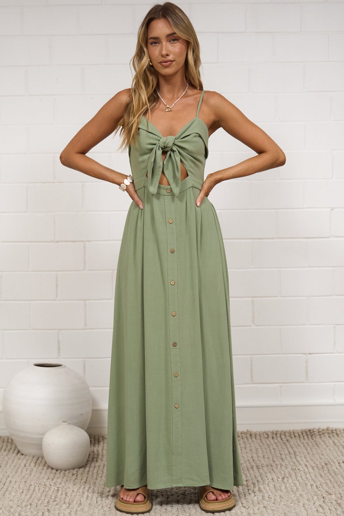 A woman modeling a sage green sleeveless maxi dress with a tie-front bodice, standing on a textured beige rug against a white brick wall.