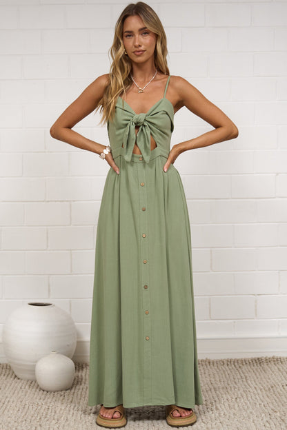 A woman modeling a sage green sleeveless maxi dress with a tie-front bodice, standing on a textured beige rug against a white brick wall.