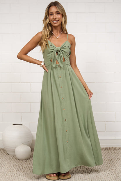 A woman modeling a sage green sleeveless maxi dress with a tie-front detail and button-down front, standing in a bright room with a white brick wall and neutral decor.