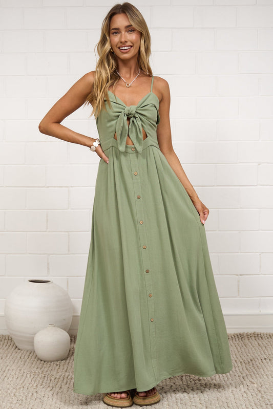A woman modeling a sage green sleeveless maxi dress with a tie-front detail and button-down front, standing in a bright room with a white brick wall and neutral decor.