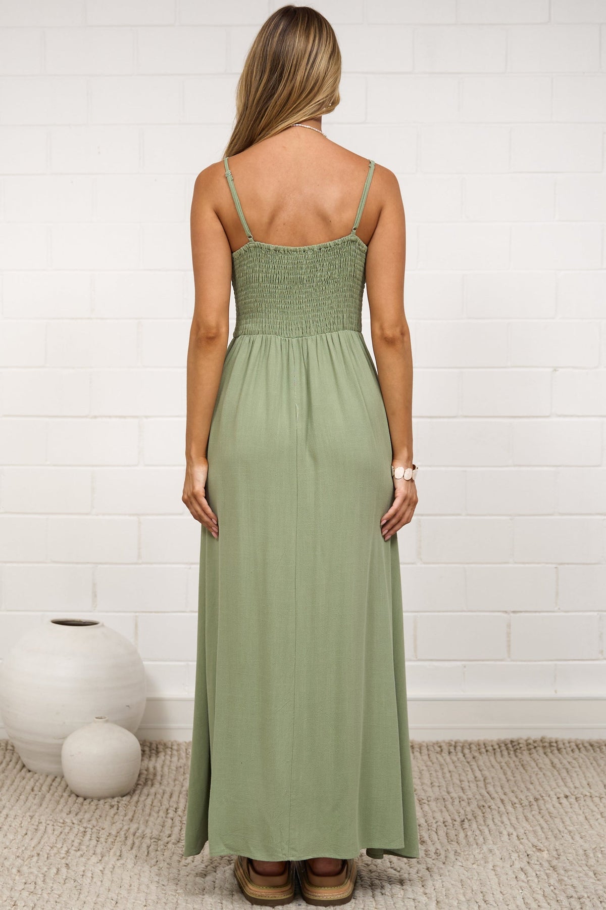 A woman stands with her back to the camera wearing a strapless sage green maxi dress in a bright, modern room with a white brick wall and neutral decor.