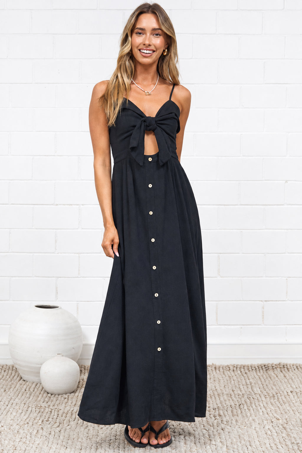 A woman modeling a black sleeveless maxi dress with a sweetheart neckline, front cutout, and a row of gold buttons down the front, standing in a minimalist room with a white brick wall and neutral rug.