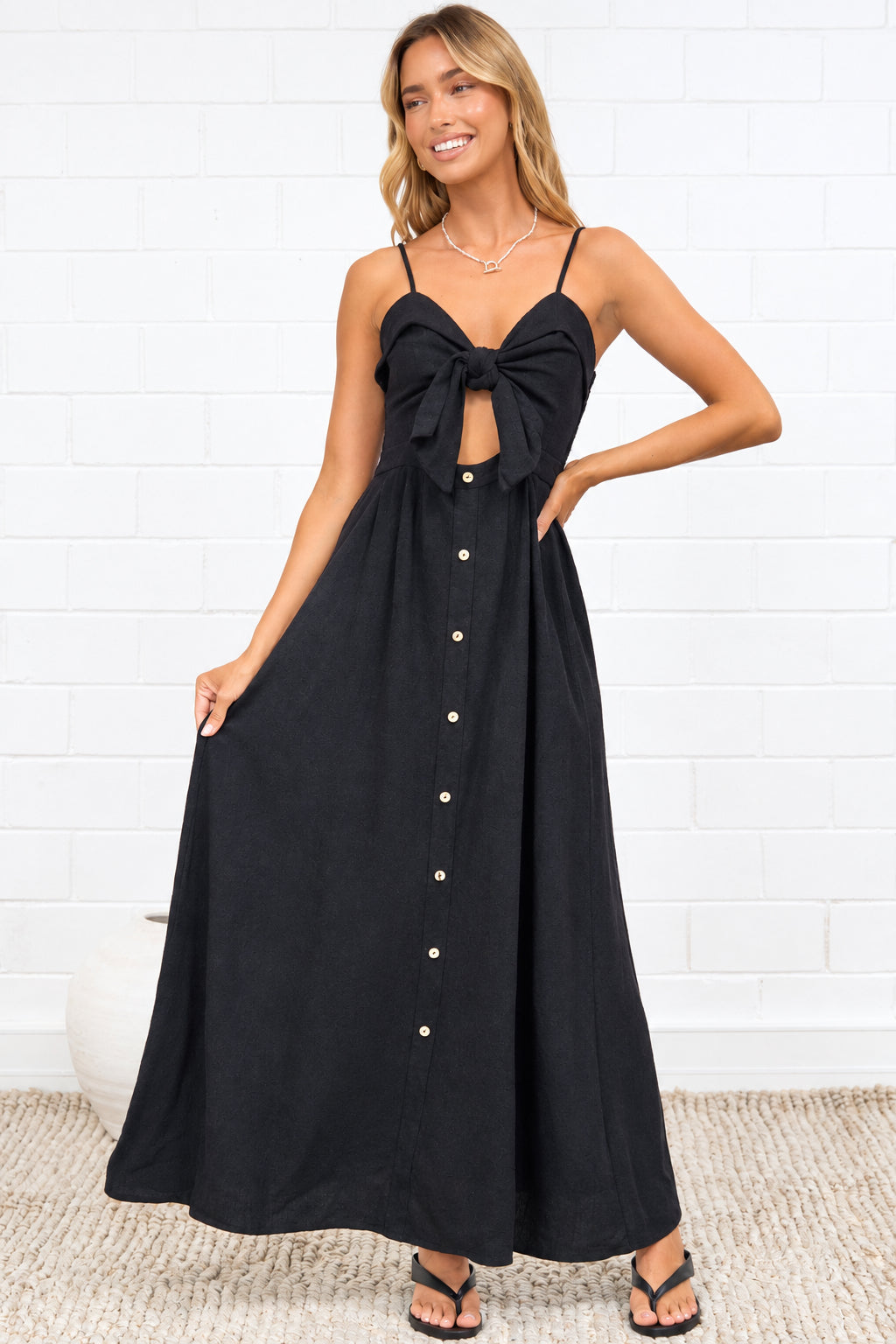A woman modeling a black maxi dress with a sweetheart neckline, a large front bow, and a button-down front, standing in a bright room with a white brick wall and a beige rug.