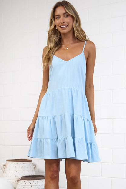 A woman modeling a light blue sleeveless tiered mini dress in a bright, modern studio setting with a white brick wall, beige rug, and decorative ceramic vases.