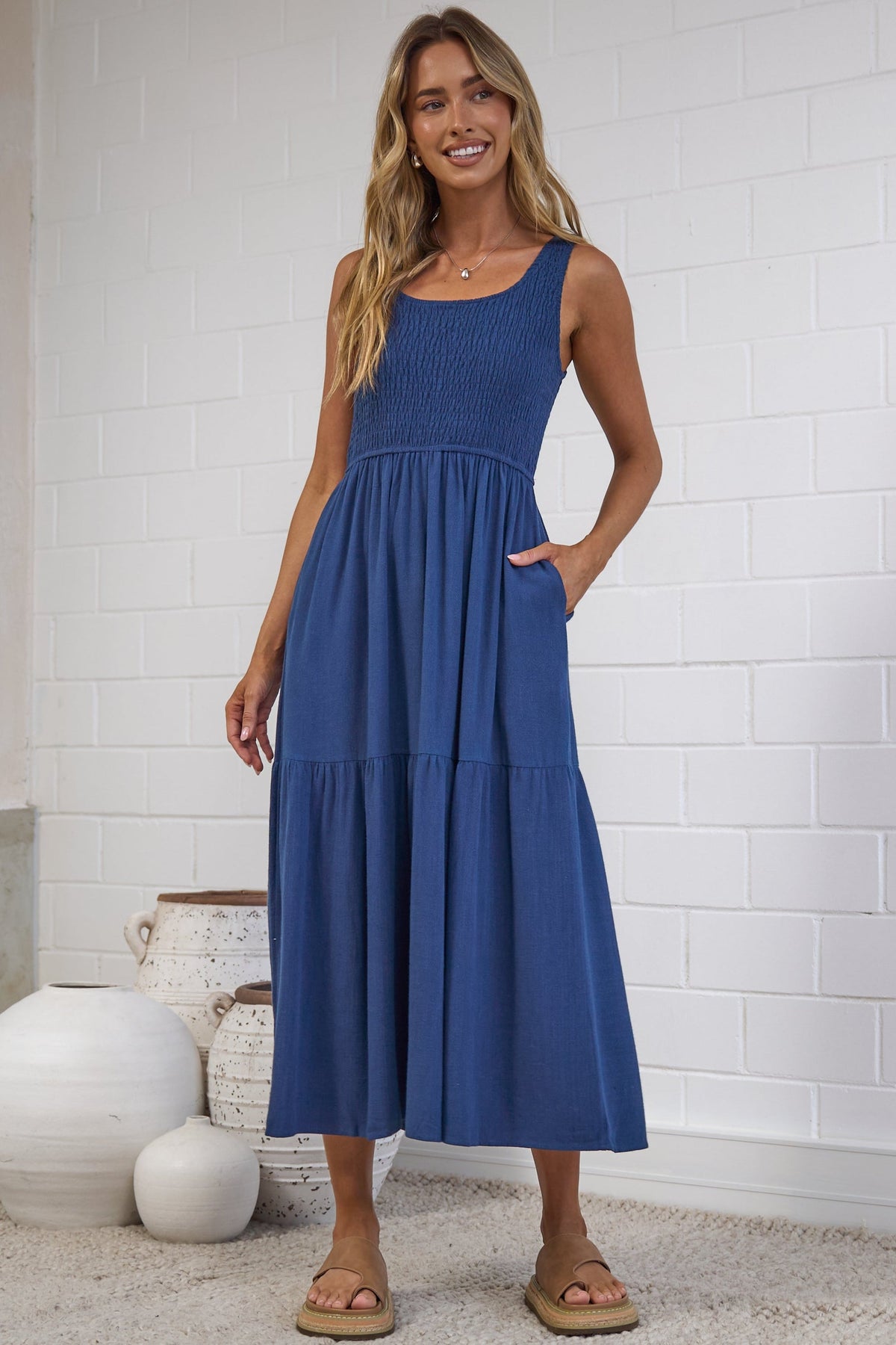 A woman modeling a sleeveless, tiered blue maxi dress with pockets, standing on a light beige rug in front of a white brick wall.