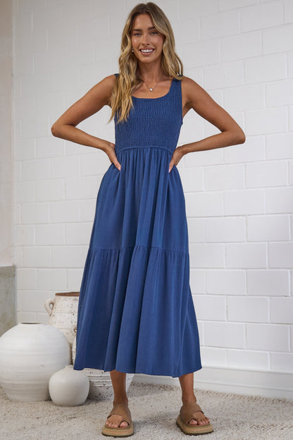 A woman modeling a sleeveless, tiered blue maxi dress in a bright, modern studio with white brick walls and neutral decor.