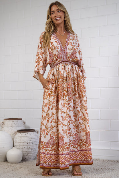 A woman modeling a bohemian-style maxi dress with a rust-orange and cream floral pattern, standing in front of a white brick wall with decorative white vases on the floor.