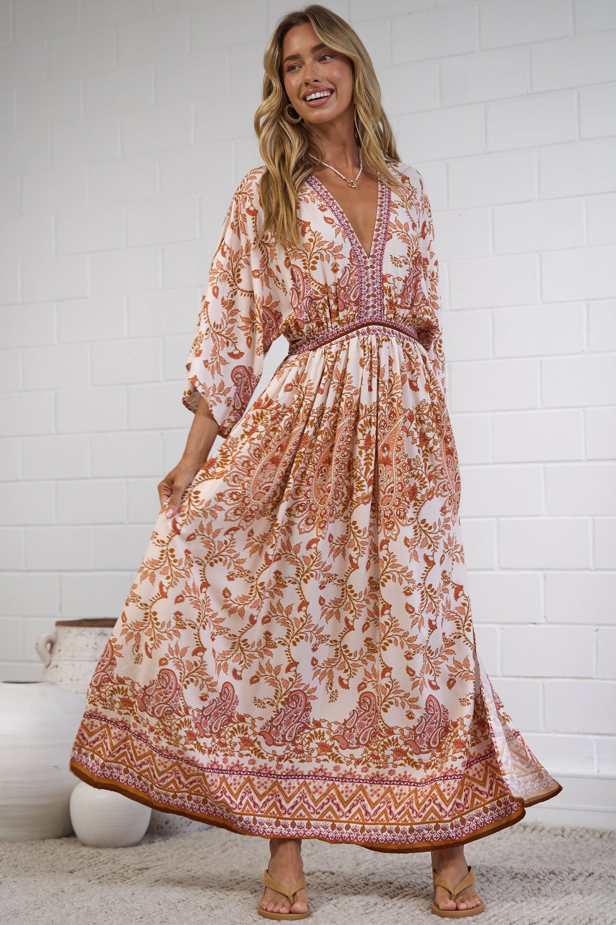 A woman modeling a bohemian-style maxi dress with a warm orange, pink, and beige floral pattern, standing in a bright room with a white brick wall and a textured rug.