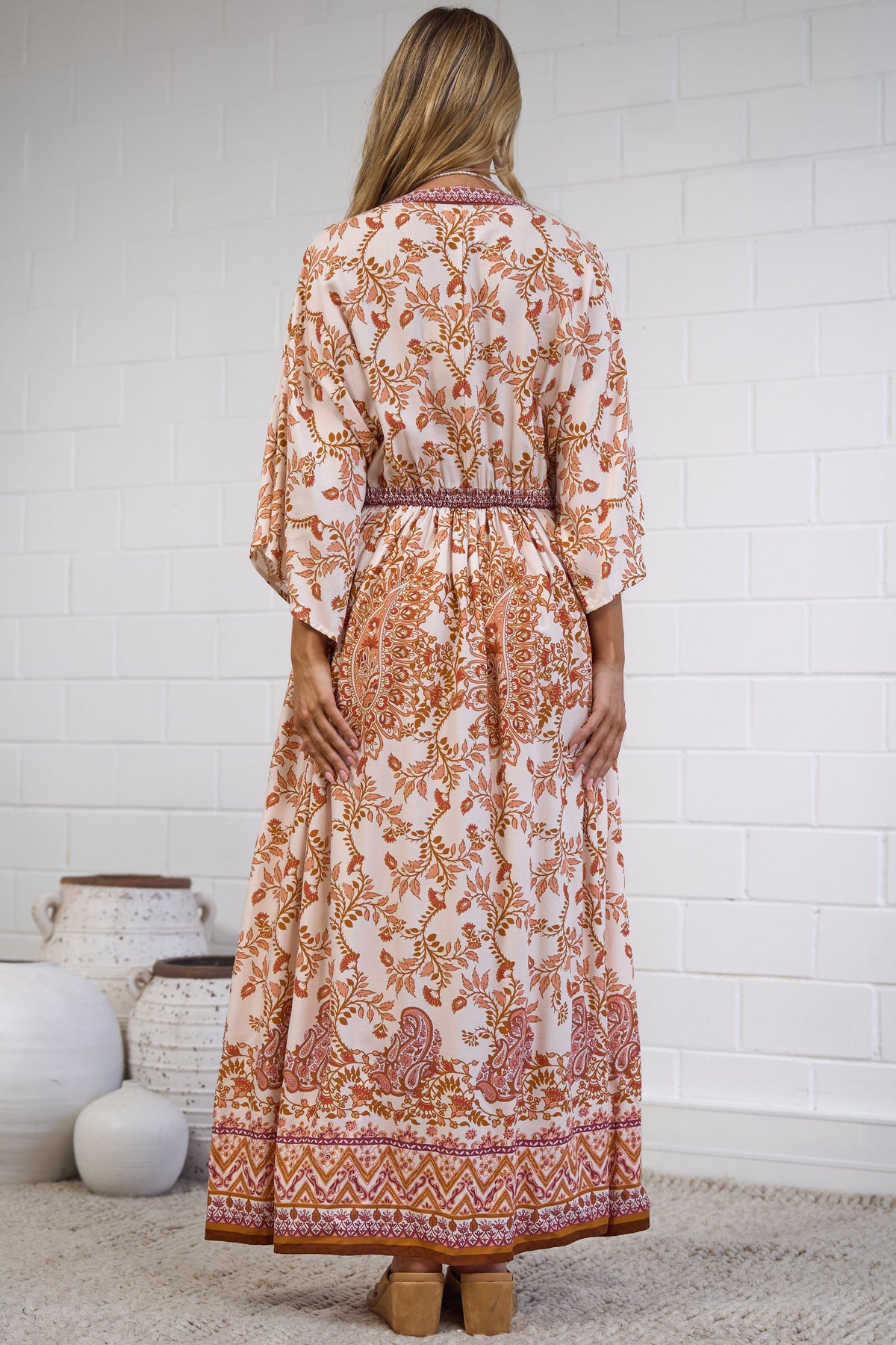 A woman stands with her back to the camera wearing a long-sleeved, ankle-length kaftan dress with an orange and brown floral pattern on a cream background. She is standing on a textured beige rug in front of a white brick wall.
