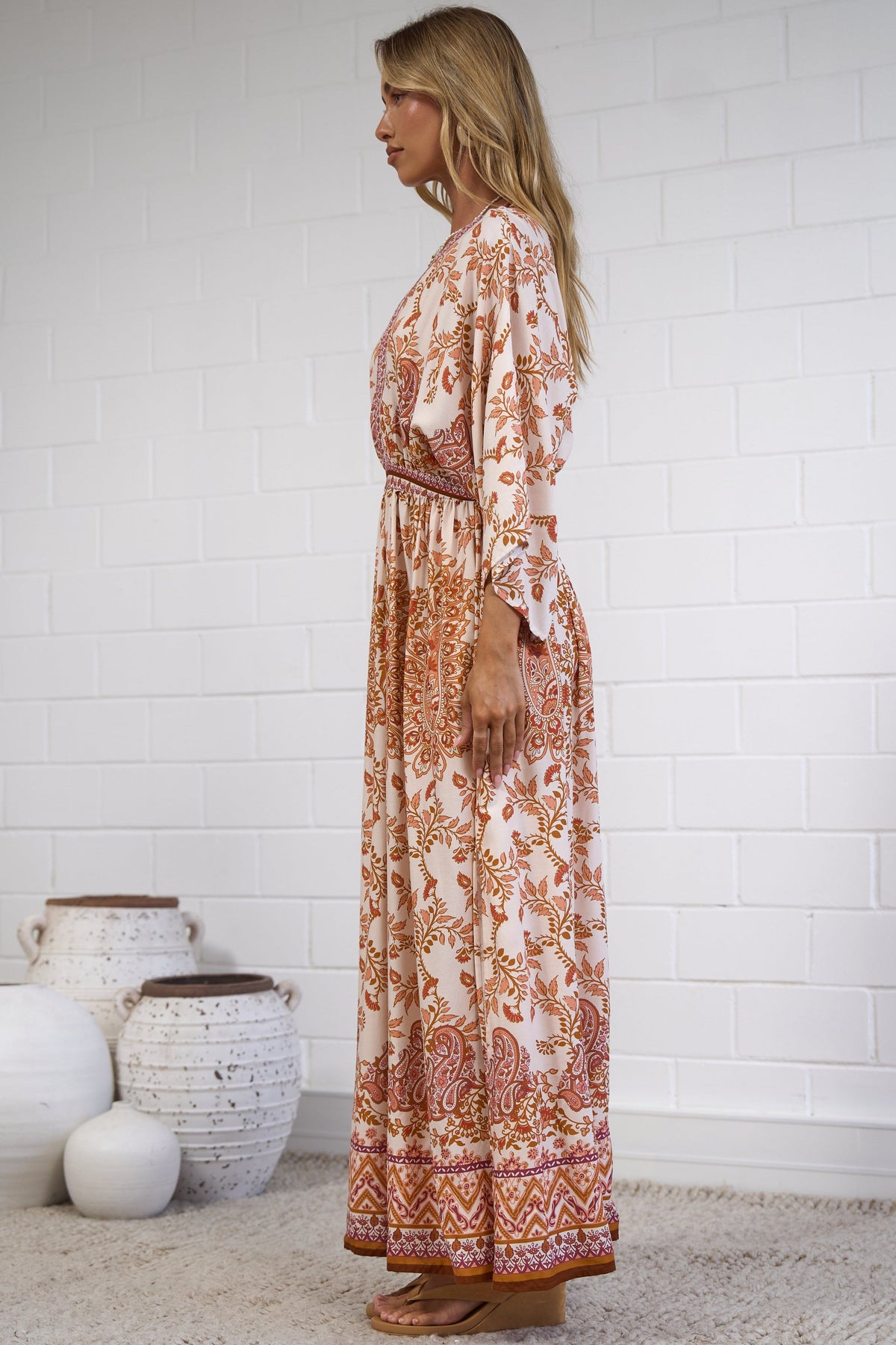 A woman stands in profile wearing a long-sleeve maxi dress with an intricate rust-orange and cream floral pattern. She is standing on a light beige woven rug against a white brick wall, with decorative white ceramic vases on the floor to her left.