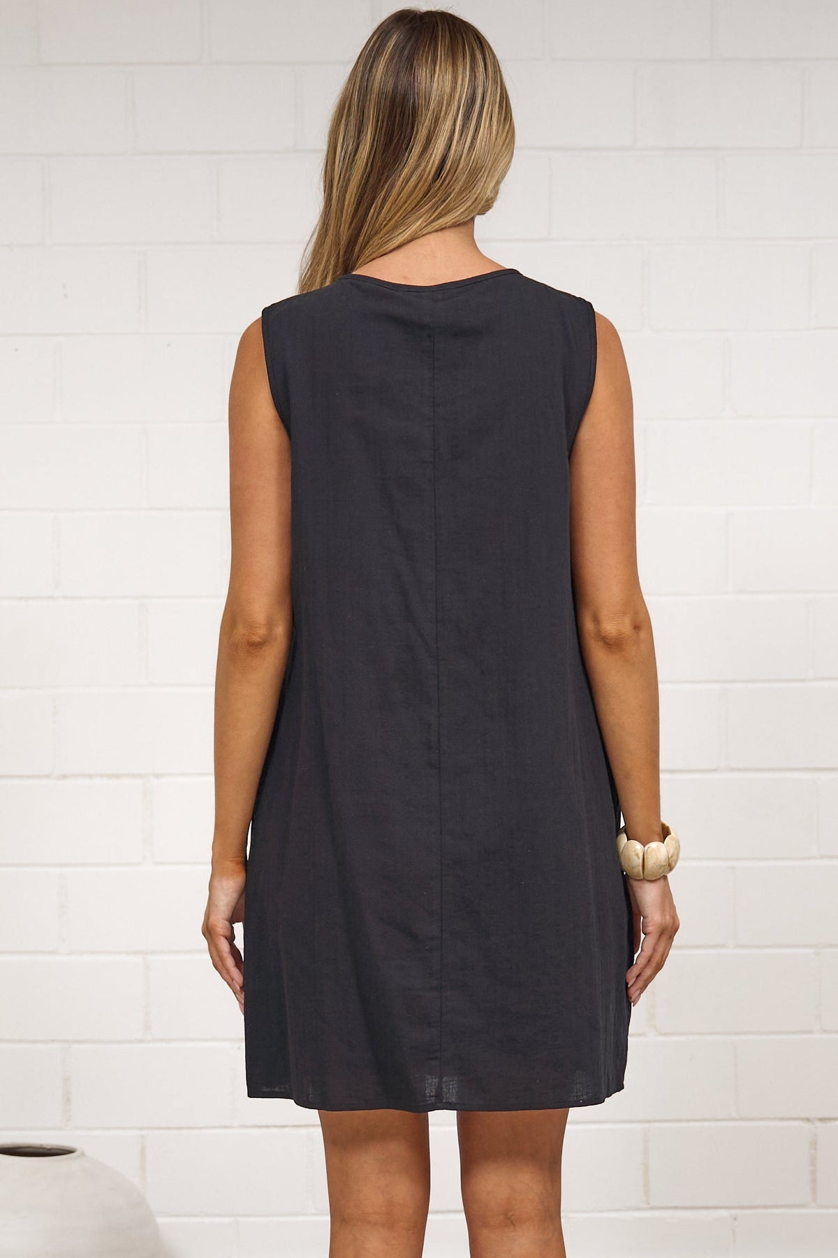 A woman stands with her back to the camera wearing a sleeveless black dress, posing in a minimalist room with a white brick wall, a beige rug, and white vases.