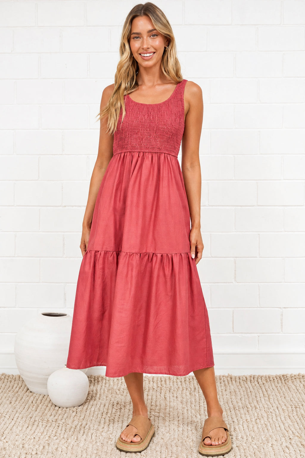 A woman modeling a sleeveless tiered midi dress in a dusty rose pink color, standing on a textured beige rug against a white brick wall.