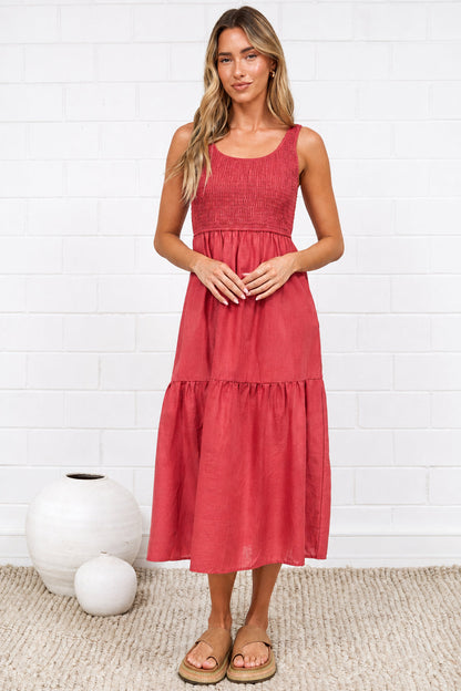 A woman stands in a bright room wearing a sleeveless, tiered midi dress in a dusty rose pink color. The room features a white brick wall, a light beige rug, and minimalist white ceramic vases.