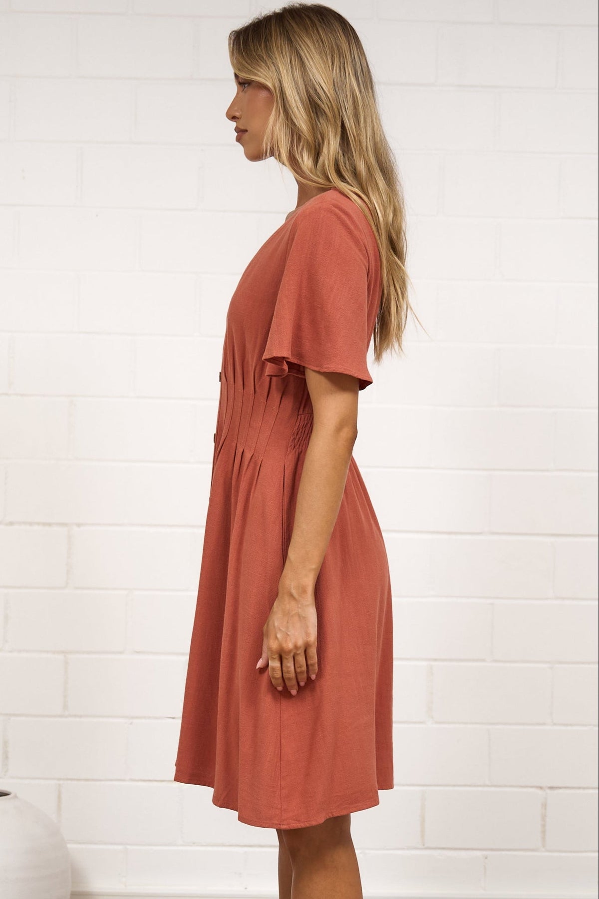 A woman stands in profile wearing a short-sleeve terracotta dress in a bright room with white brick walls, a beige rug, and minimal decor.