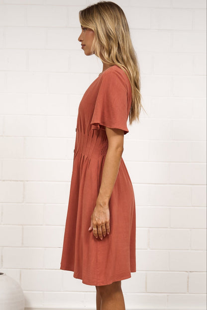 A woman stands in profile wearing a short-sleeve terracotta dress in a bright room with white brick walls, a beige rug, and minimal decor.