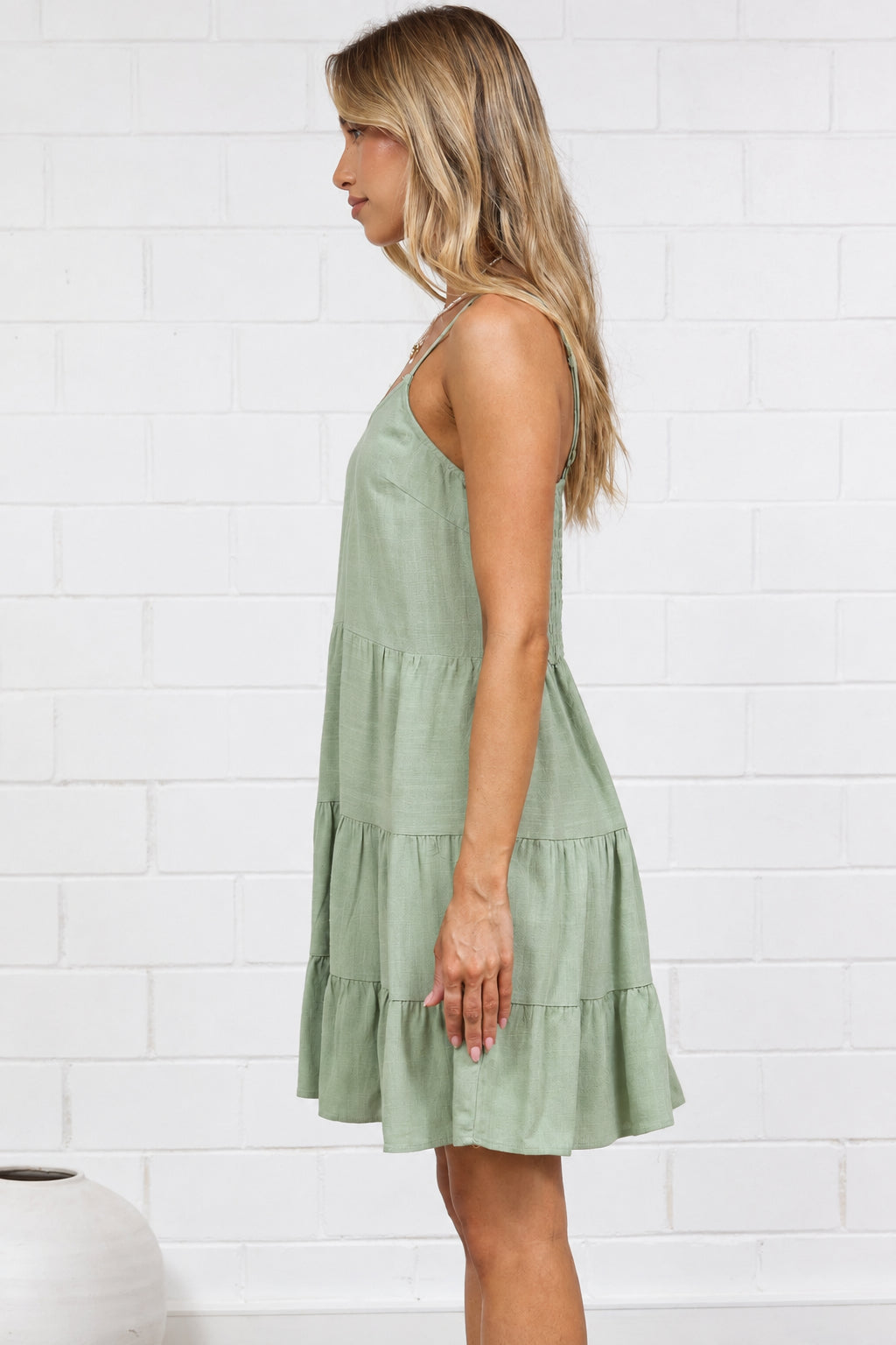 A woman stands in profile wearing a sleeveless olive green tiered dress, standing on a light beige jute rug against a white brick wall.